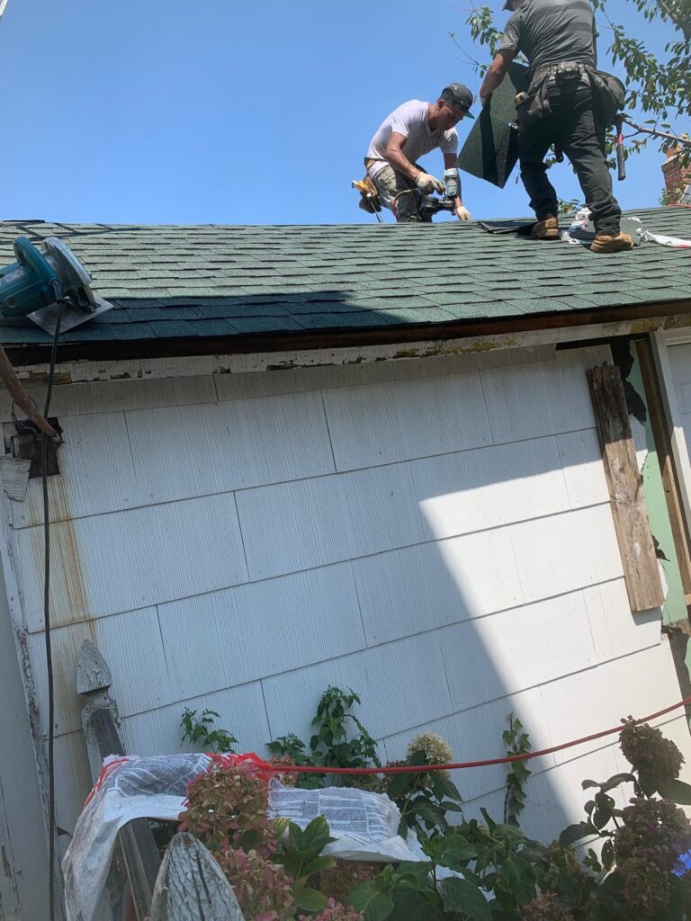 Workers repairing a roof under blue skies, using tools and holding roofing material; plants nearby Bronx, NY